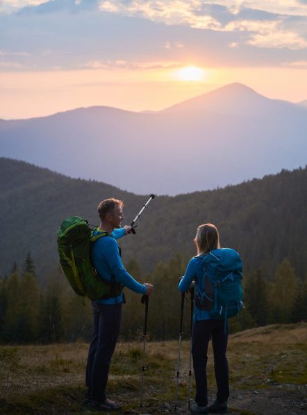 Back view of young man with tourist backpack holding up trekking stick in his hand, pointing to sunset over the mountain peaks to his wife in evening. Concept of travelling, hiking and active leisure.